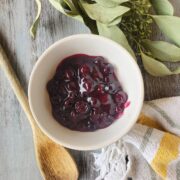 White bowl containing blueberry sauce with greenery, a wooden spoon, and a kitchen towel surrounding it